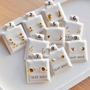 May include: A collection of gold-colored stud earrings displayed on white earring cards. The earrings feature various celestial designs, including stars, moons, planets, a rocket, and an astronaut. Each card is labeled "10 KT. GOLD."
