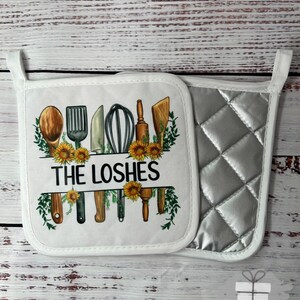 May include: White pot holder set with a silver quilted pot holder. The white pot holder has a design of kitchen utensils, sunflowers, and the text "THE LOSHES". The set is trimmed with white binding.