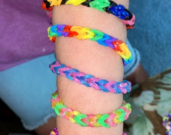 May include: A person's arm wearing a stack of colorful rainbow loom bracelets. The bracelets are made of rubber bands and are in various colors, including pink, purple, blue, green, yellow, orange, and red.