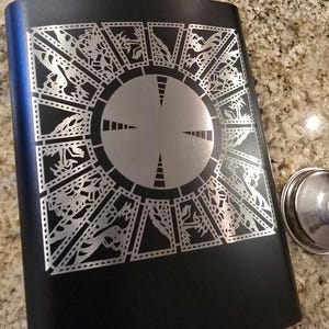 May include: Black metal flask with a silver engraved design of a puzzle box with a circular center.