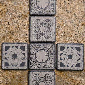 May include: Set of six black slate coasters with a white design of a complex puzzle box. The coasters are arranged in a vertical stack of three, with two coasters on either side.