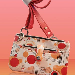 May include: A white crossbody bag with a red strap and a geometric pattern of red, orange, and black circles and lines. The bag has two zippered compartments and a snap closure.
