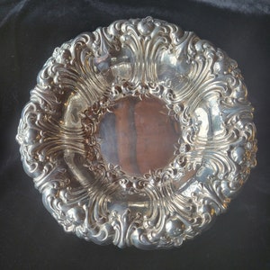 May include: A silver serving bowl with an ornate, embossed design. The bowl has a round shape and a wide, shallow rim.