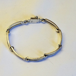 May include: A silver bracelet with a simple, geometric design. The bracelet is made of linked, flat, rectangular pieces with rounded edges. The clasp is marked with the number "925", indicating that the bracelet is made of sterling silver.