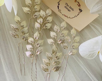 Könnte beinhalten: Fünf goldene Haarspangen mit einem zarten Zweigdesign. Die Spangen sind auf einem weißen Tüllstoffhintergrund angeordnet. Der Text "The little things" ist auf einer Karte im Hintergrund sichtbar.