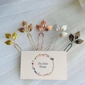 May include: Five gold leaf hair clips in different colors: gold, rose gold, silver, bronze, and green. The clips are arranged in a row on a white background. The text "The little things" is printed on a card below the clips.