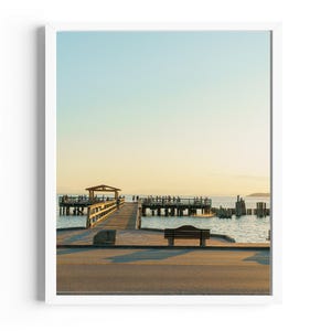 以下が含まれることがあります： 木製の桟橋が水の中に伸びており、人々が立っています。桟橋は風化した木でできており、端に小さな木製の小屋があります。水は穏やかで青く、空は薄い青で、雲が少しあります。木製のベンチが手前にあります。