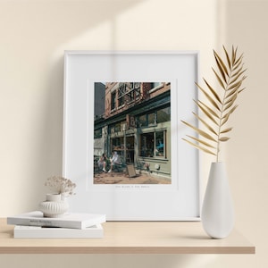 Op de afbeelding: Een ingelijste prent van een caféscène, met een groen en bakstenen gebouw met zitplaatsen buiten. De prent staat op een houten plank met een witte vaas, gedroogde bloemen en boeken. De tekst "The Birds & The Beets" is zichtbaar.