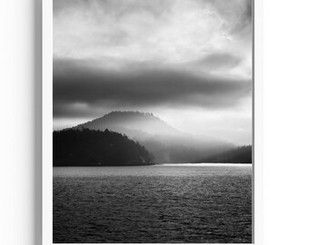 May include: A black and white photograph of a misty mountain range with a lake in the foreground. The sky is cloudy and the sun is shining through the clouds.