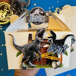 Puede incluir: Una caja blanca con forma de frontón con temática de Parque Jurásico. La caja presenta un gráfico de dinosaurio, el texto "James" y las palabras "Caution" en cinta amarilla. La caja está decorada con siluetas de dinosaurios y huevos.