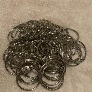 May include: A large collection of silver-colored metal loose-leaf binder rings. Each circular ring features a hinged mechanism that allows it to open and close securely. The rings are made of polished steel and are designed for organizing papers.