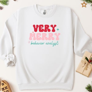 May include: White sweatshirt with a red, pink, and teal graphic that says "VERY MERRY behavior analyst".
