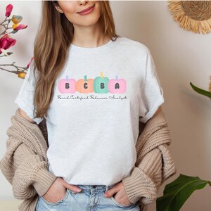 Puede incluir: Una camiseta blanca con un gráfico de cuatro calabazas con las letras "BCBA" en ellas. El texto "Board Certified Behavior Analyst" está escrito debajo de las calabazas.