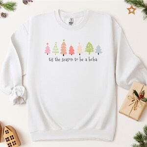 May include: White sweatshirt with a festive design of colorful Christmas trees and the text "tis the season to be a beba".