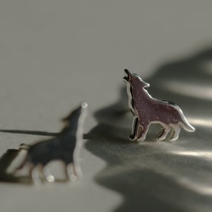 May include: Two silver-colored wolf-shaped stud earrings. One earring is in focus, depicting a howling wolf. The other earring is slightly out of focus, with a shadow cast on the surface. The earrings are likely made of metal.