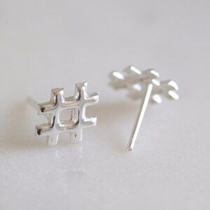 May include: A pair of silver-colored hashtag stud earrings. The earrings are in the shape of a hashtag symbol, with a post on the back for insertion into the ear. The earrings are displayed on a white surface.