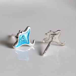 May include: A pair of silver stud earrings featuring a blue enamel manta ray design. The earrings are shown on a white surface.