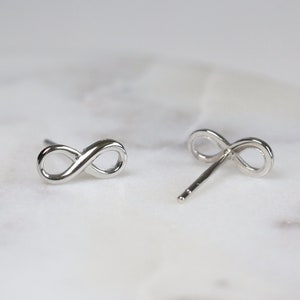 May include: A pair of silver infinity symbol stud earrings. The earrings are on a white marble surface.