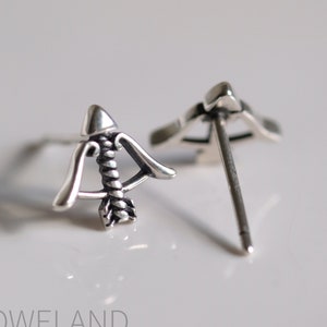 May include: A pair of silver-colored stud earrings shaped like a bow and arrow. The arrow is pointing towards the right, and the bow has a curved design. The earrings are on a white surface.