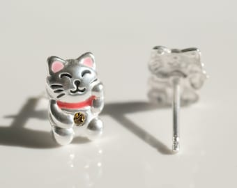 May include: A pair of silver stud earrings featuring a small, smiling, pink-cheeked, white cat with a red collar and a gold coin on its chest. The cat is in a sitting position with its paws raised.