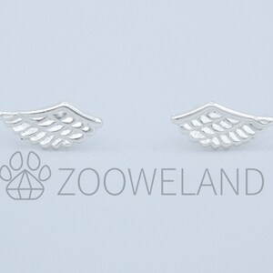 May include: A pair of silver-coloured wing-shaped stud earrings. Each earring has a detailed feather design, with a curved top and parallel lines representing feathers. The earrings are presented against a light background.