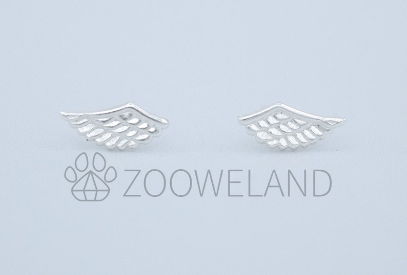May include: A pair of silver-coloured wing-shaped stud earrings. Each earring has a detailed feather design, with a curved top and parallel lines representing feathers. The earrings are presented against a light background.