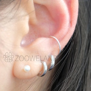 May include: Close-up of an ear with multiple earrings. A small, white pearl stud is in the lobe, and two silver hoop earrings and a silver ring earring are visible. The ear is against a blurred background.