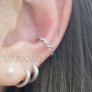 May include: A silver-colored, twisted helix earring is shown in the upper ear. The earring is a small, continuous hoop with a braided design. The image also shows two additional silver hoop earrings and a small pearl stud in the lower ear. The brand name "ZOOWELAND" is visible.