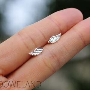 May include: A pair of small, silver wing-shaped stud earrings. The earrings are displayed on a person's hand, with a blurred green background. The earrings have detailed feather designs.