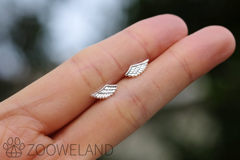 May include: A pair of small, silver wing-shaped stud earrings. The earrings are displayed on a person's hand, with a blurred green background. The earrings have detailed feather designs.