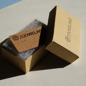 May include: A light brown cardboard box, open to reveal a smaller tag inside, both branded with the "ZOOWELAND" logo. The box contains white packing material. The tag has two small holes. The box is rectangular.