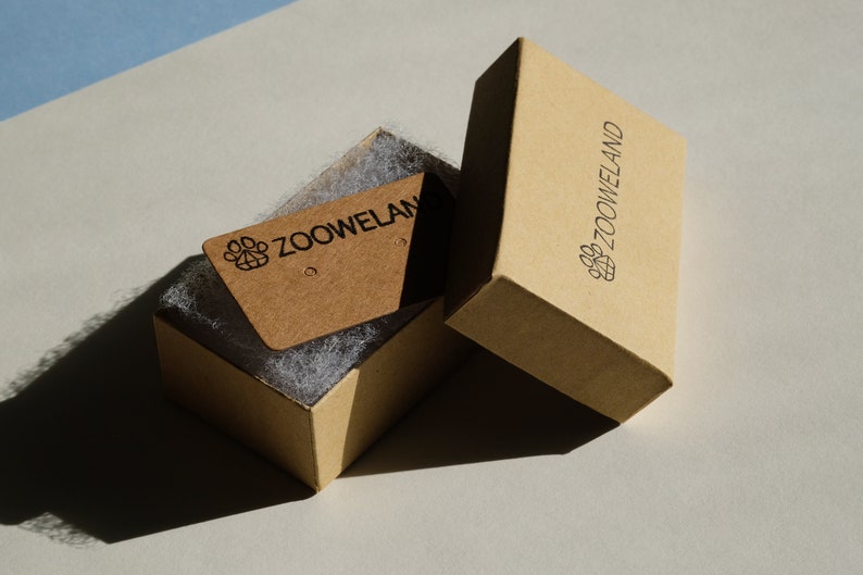 May include: A light brown cardboard box, open to reveal a smaller tag inside, both branded with the "ZOOWELAND" logo. The box contains white packing material. The tag has two small holes. The box is rectangular.