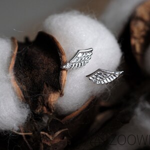 May include: A pair of silver wing-shaped stud earrings. The earrings are set against a backdrop of white cotton and brown seed pods. The earrings feature detailed feather-like designs.