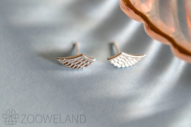 May include: A pair of silver-coloured wing-shaped stud earrings. Each earring has a detailed feather design. The earrings are displayed on a light blue, silky fabric.