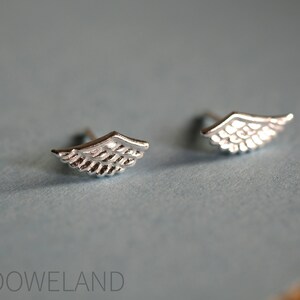 May include: A pair of small, silver-coloured wing-shaped stud earrings. The earrings feature feather-like patterns and are displayed on a light blue surface. The earrings are a jewellery item.