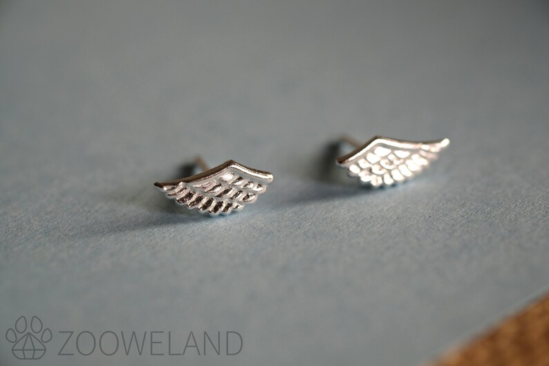 May include: A pair of small, silver-coloured wing-shaped stud earrings. The earrings feature feather-like patterns and are displayed on a light blue surface. The earrings are a jewellery item.