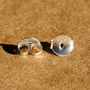May include: Two silver earring backs, one with a butterfly shape and the other with a post and the number "925" stamped on it. The earring backs are on a textured, brown surface.