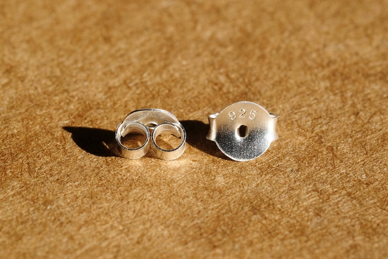 May include: Two silver earring backs, one with a butterfly shape and the other with a post and the number "925" stamped on it. The earring backs are on a textured, brown surface.