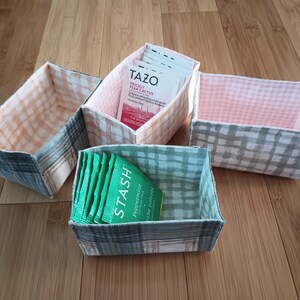 May include: Four fabric storage boxes with a checkered pattern. Two boxes are pink and white, one is light blue and white, and one is gray and white. The gray and white box contains a pack of green tea bags labeled "STASH Peppermint" tea bags.