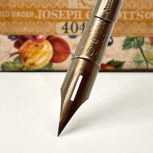May include: Close-up of a vintage brass pen nib, model 404, with the text "JOSEPH GILLOTT'S". The nib is pointed and has a slit down the center. The background features a decorative box with floral and fruit illustrations.