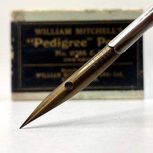 May include: A gold-colored metal nib for a fountain pen. The nib is labeled "Pedigree" and "C.C.C.P." The nib is sitting on a white surface with a black box in the background.