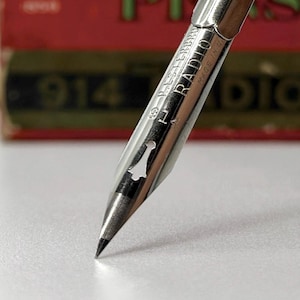 May include: Close-up of a silver-colored pen nib with the text "ESTERBROOK RADIO MADE IN U.S.A." The nib is pointed downward, with a fine tip. The background is a blurred red and green.