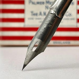 May include: Close-up of a vintage metal calligraphy pen nib. The nib is silver-toned with the text "PALMER MET No 5" etched on the side. The tip is finely pointed. The background features a red and white striped pattern.