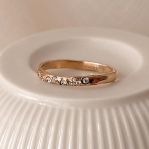 Op de afbeelding: Een goudkleurige ring met een getextureerde band en kleine, heldere edelstenen. De ring ligt op een wit, geribbeld oppervlak, met een zachte, neutrale achtergrond. Het ontwerp is eenvoudig en elegant, geschikt voor dagelijks gebruik of speciale gelegenheden.