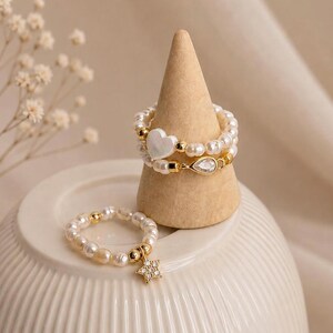 Könnte beinhalten: Eine Auswahl an Perlenringen. Ein Ring hat einen goldenen Sternanhänger mit klaren Kristallen. Zwei Ringe werden auf einem Holzkegel präsentiert, einer mit einem herzförmigen Anhänger und der andere mit einem tropfenförmigen Anhänger. Die Ringe haben goldene Akzente.