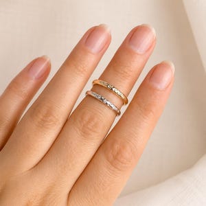 Peut inclure: Deux bagues fines, l'une dorée et l'autre argentée, chacune ornée de petites pierres scintillantes. Les bagues sont présentées sur un doigt, mettant en valeur leur design simple mais élégant. La bague dorée est au-dessus, et l'argentée en dessous.