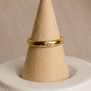 Puede incluir: Un anillo dorado con una pequeña gema azul claro engastada en la banda. El anillo se exhibe sobre un soporte de madera en forma de cono, sobre un fondo neutro. El anillo tiene un diseño de banda simple.