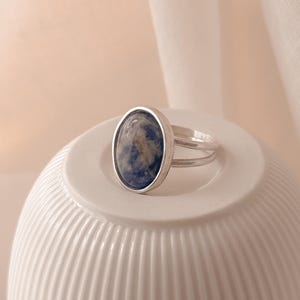 Könnte beinhalten: Ein silberner Ring mit einem ovalen, blauen Edelstein. Der Ring hat ein Doppelband-Design und liegt auf einer weißen, geriffelten Oberfläche. Der Edelstein hat blaue und braune Farbtöne.