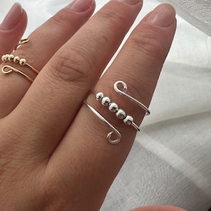 May include: Close-up of three rings. The main ring is silver with four small, silver beads and a curved design. A gold ring with small gold beads is visible in the background. The rings are on a finger.