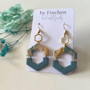 Könnte beinhalten: Handgefertigte Ohrringe mit geometrischem Design in Teal und Gold. Die Ohrringe haben eine sechseckige Form mit goldfarbenen Metallakzenten. Sie werden auf einer weißen Karte mit dem Text "by Finchen hand crafted jewelry" präsentiert.